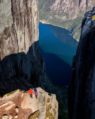 İki maceraperest bir uçurumun kenarında duruyor, Lysefjord 'un çarpıcı mavi sularına bakıyor, berrak bir gökyüzünün altında yükselen kayalarla çevrili. Kjerag Kjeragbolten Lysefjord Norveç
