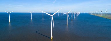 Uçsuz bucaksız rüzgâr türbinleri temiz enerji üretirken berrak mavi gökyüzünün altındaki sakin suların üzerine gölgeler düşürüyor. Hollanda 'da bir yel değirmeni parkında insansız hava aracı görüntüsü