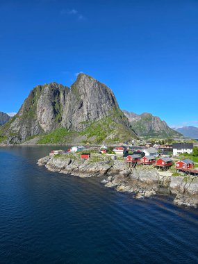 Açık mavi gökyüzünün altında yüksek dağlarla çevrili kıyı şeridi boyunca uzanan kırmızı kulübeleri olan pitoresk bir köy. Lofoten Norveç 