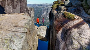 İki maceracı kayalık bir çıkıntıda dikkatle yürürken, Kjeragbolten mavi bir fiyort üzerindeki uçurumlar arasında dramatik bir şekilde dengede duruyor. Kjerag Kjeragbolten Lysefjord Norveç 'te bir çift kadın ve erkek