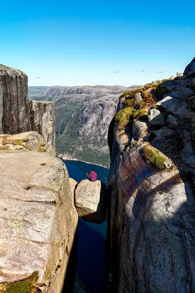 stock image An adventurous soul perches on a precarious rock, surrounded by breathtaking fjords and towering cliffs under a clear blue sky. Kjerag Kjeragbolten Lysefjord Norway