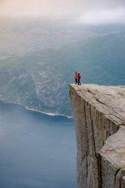 Kırmızı ceketli iki yürüyüşçü, aşağıdaki fiyordun çarpıcı manzarasıyla dramatik bir uçurumun kenarında güven içinde dikiliyorlar. Önceki Norveç