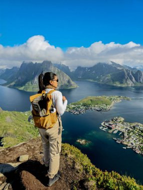 Bir insan, engebeli tepeler ve parlak mavi gökyüzünün altındaki sakin sularla çevrili bir dağın tepesinden nefes kesici fiyort görüntülerine hayrandır. Reinebringen Lofoten Norveç 