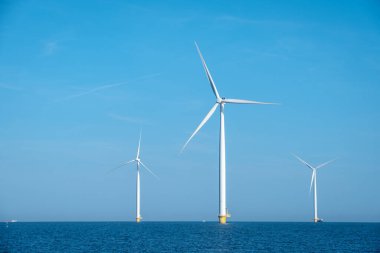 Uzun rüzgar türbinleri açık mavi gökyüzüne çarpıcı bir siluet oluşturarak Hollanda kıyı şeridinin hafif dalgaları ve meltemlerinden gelen enerjiyi kullanırlar. Kıyıdaki yel değirmeni parkı, Hollanda.