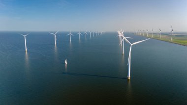 Bir yelkenlinin açık mavi gökyüzüne karşı yükselen rüzgar türbinleri arasında huzur içinde süzülmesiyle sakin bir sabah başlıyor. Hollanda 'daki yel değirmeni parkı. Ijsselmeer Gölü.