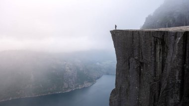 Yalnız bir figür Preikestolen 'in kenarında duruyor, şafak sökerken sisli Lysefjord' a bakıyor, dramatik uçurumlara ve sakin sulara ruhani bir ışık tutuyor.. 