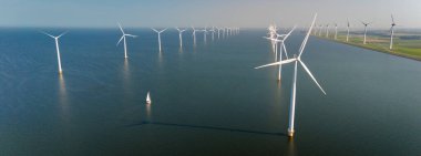 Hollanda açıklarında sakin bir yel değirmeni parkı manzarası, sabah gökyüzüne karşı zarif dönen uzun türbinler, Hollanda sularında sessizce süzülen küçük bir yelkenli.