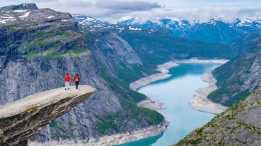 İki dağcı, uzaktaki karlı dağlarla kaplı Norveç fiyortlarına tepeden bakan kayalık bir uçurumda dururlar. Trolltunga, Norveç,