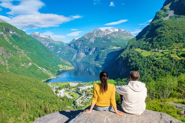 Bir çift, yeşil dağlar ve açık mavi gökyüzü ile çevrili, göz kamaştırıcı bir Norveç fiyorduna tepeden bakan kayalık bir çıkıntıda oturuyor. Geiranger Fjord Norveç 'te çeşitli kadın ve erkekler var.