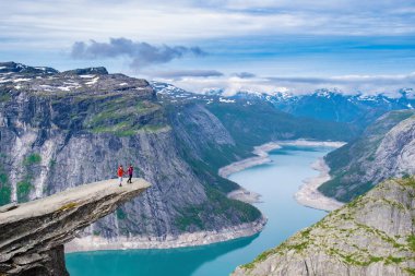 Norveç 'teki Trolltunga uçurumunun kenarında duran bir çift fiyortun çarpıcı mavi sularına bakıyor. Norveç 'teki Trolltunga Dağı' nın kıyısında bir çift kadın ve erkek.