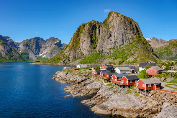 stock image red cabins perched on the rocky shore of a fjord in Norway, surrounded by towering mountains and a clear blue sky. Hamnoy fishing village on Lofoten Islands, Norway with red rorbu houses