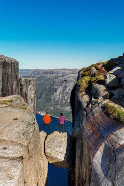 Canlı ceketli iki kaşif kayalık bir çıkıntıda dengede duruyor, açık mavi gökyüzünün altındaki nefes kesici fiyorda hayret ediyorlar. Kjerag Kjeragbolten Lysefjord Norveç