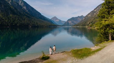 Bir çift Plansee Gölü 'nün berrak sularının kenarında, görkemli dağlarla çevrili huzurlu bir yürüyüşün tadını çıkarıyor. Huzurlu manzara, doğa ve sükunetin mükemmel bir uyumunu yansıtır..
