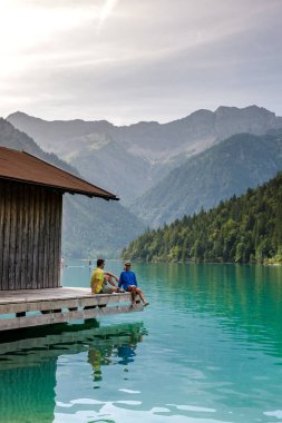 A couple of man and woman enjoy a tranquil afternoon by the emerald waters of Plansee Austria, surrounded by breathtaking mountains and lush greenery, creating a peaceful retreat in nature's embrace. clipart