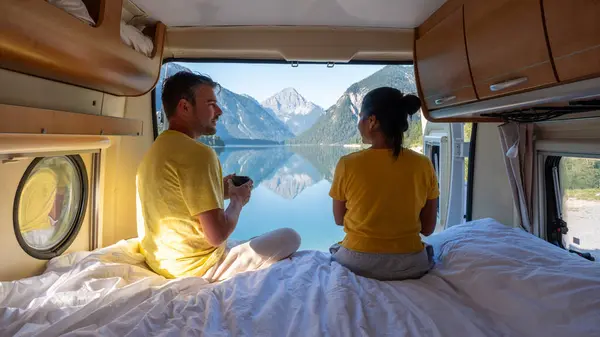 stock image A peaceful morning is unfolding as a couple relaxes in their camper van by Plansee Lake Austria. Surrounded by majestic mountains, they savor hot drinks while enjoying the serene landscape.