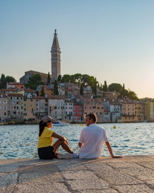 Limanın kenarında oturan iki kişi, Hırvatistan 'ın Rovinj kentinin canlı binalarına bakıyor. Batan güneş altın gibi parlıyor, tarihi çan kulesini ve dingin suları aydınlatıyor..