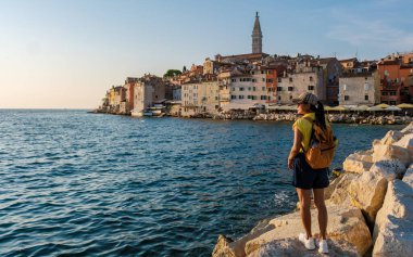 Gün batımında, Rovinj 'in sakin sularından büyülenmiş bir gezgin kayalık sahilde duruyor. Tarihi binalar sönen ışıkta parlıyor, macera ve huzur hissi uyandırıyor..