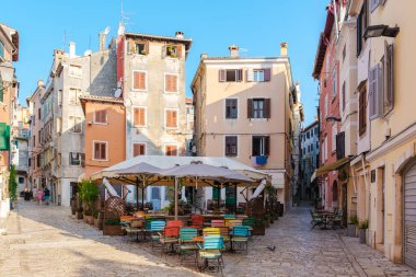 Rovinj 'deki canlı bir açık kafe, müşterileri geniş bir gölgelik altında canlı oturmanın keyfini çıkarmaya davet ediyor. Bu kıyı kasabasının büyüleyici caddeleri sıcak güneş ışığının tadını çıkarıyor, davetkar bir atmosfer yaratıyor..