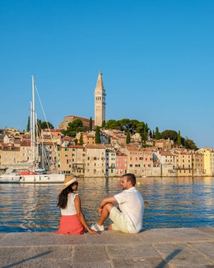 Bir çift, Rovinj 'in dingin suyuna tepeden bakan taş bir rıhtımda oturmaktan keyif alıyor. Büyüleyici sahil kasabası renkli binalarla ve arka planda tarihi bir çan kulesiyle övünüyor..