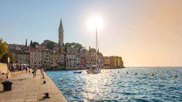 stock image As the sun sets over Rovinj, its golden rays enhance the vibrant, colorful buildings along the waterfront. A sailboat gently rocks on the serene waters, inviting evening tranquility.