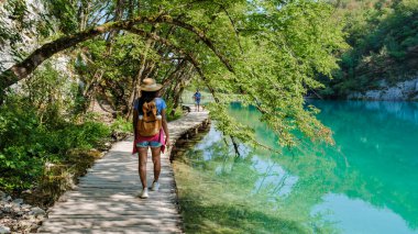 Bereketli yeşilliklerin ortasında gezginler Plitvice Gölleri Ulusal Parkı 'nın ahşap geçitlerinde turkuaz suların ve berrak bir gökyüzü altında canlı bir doğanın huzurlu güzelliği içinde gezinirler..