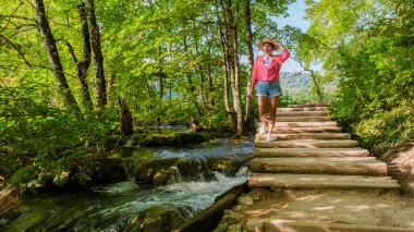 Yeşillik içinde gezen bir gezgin, Plitvice Gölleri 'nin dingin güzelliğinin tadını çıkarır. Güneş ağaçların arasından süzülür, akan su ile ahşap yollar boyunca canlı doğayı aydınlatır..