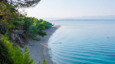 Güneş ışığı, Brac Adası 'nın sakin kıyılarını süslüyor. Yumuşak dalgaların kumlu sahili okşadığı, sakin yürüyüşleri davet ettiği kıyıları. Yeşillikler kıyı şeridini çevreleyerek, ziyaretçiler için huzurlu bir geri çekilme oluşturuyor..