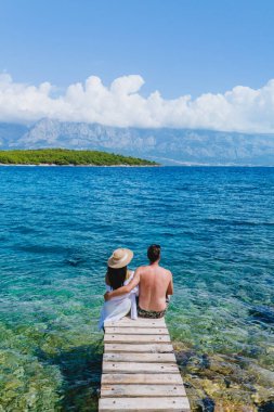 Bir çift, Brac Adası 'nın sakin turkuaz sularını seyrederek, bir rustik ahşap iskelede yan yana oturur. Arkaplanda yemyeşil tepeler yükseliyor, güzel mavi bir gökyüzü tarafından çerçevelenmiş..