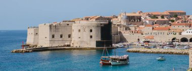 Ziyaretçiler Dubrovnik Hırvatistan 'ın ikonik taş duvarları boyunca yürürken, parlak mavi gökyüzünün altında renkli tekneler yavaşça sallanıyor.