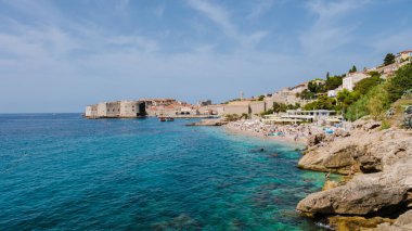 Ziyaretçiler, Hırvatistan 'ın Dubrovnik kentinde turkuaz sularının canlı uçurumlar ve tarihi yapılarla buluştuğu güzel plaj boyunca güneşlenerek, herkes için pitoresk bir sahil kaçışı yaratıyor..