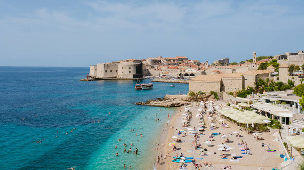 Посетители греются под теплым солнцем на песчаном пляже Дубровника, наслаждаясь кристально чистой водой и яркой исторической архитектурой вдоль берега. Очаровательная лодка мирно дрейфует в гавани.
