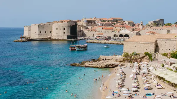 Ziyaretçiler Dubrovniks plajının yumuşak kumları ve canlı mavi sularının keyfini çıkarırken, tarihi yapılar arka planda ihtişamlı bir şekilde durarak Hırvatistan 'a güneşin altında zengin bir kültür mirası sunuyor..