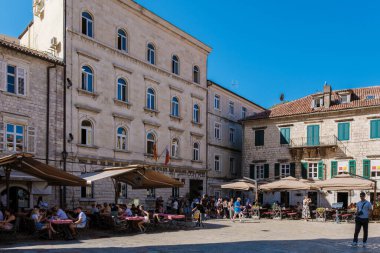 Kotor Montenegro 24 Eylül 2024, Ziyaretçiler açık mavi gökyüzü altında büyüleyici mimari ile çevrili Kotors tarihi meydanında bulunan büyüleyici açık hava kafelerindeki canlı atmosferin keyfini çıkarıyorlar..