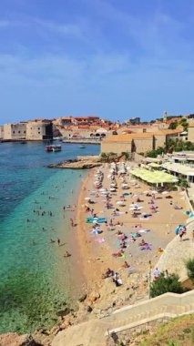 Temiz suların altın kumlarla buluştuğu ve güneşlenenlerin mavi gökyüzünün altında keyifli ve güneşli bir günün tadını çıkardıkları Hırvatistan 'ın büyüleyici Dubrovnik şehrinde büyüleyici bir plaj sahnesi.