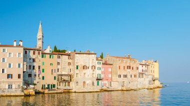Hırvatistan 'ın Rovinj kentinin sakin rıhtımında uzanan renkli eski binalar, sakin denize canlı renkler yansıtıyor. Parlak gökyüzü ve tarihi mimari resim gibi bir yaz günü atmosferi yaratır..
