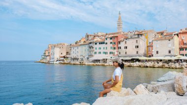 Rovinj 'in sakin sularını seyreden bir kadın kayalık sahilde huzur içinde oturuyor. Arkasından renkli binalar yükseliyor, bu güzel sahil kasabasının cazibesini ve güzelliğini yansıtıyor..