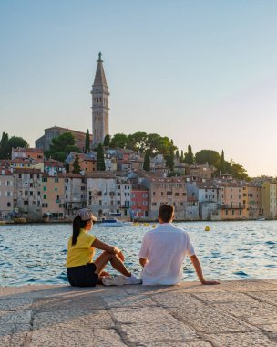 Güneş Rovinj 'in üzerinde batarken, bir çift su kenarında huzurlu bir anın tadını çıkarıyor, tuhaf pastel binalar ve tarihi bir çan kulesi ile çevrili, mükemmel bir romantik atmosfer yaratıyor..