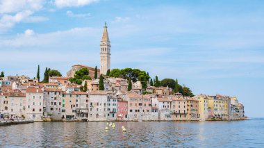 In Rovinj, Croatia, charming pastel-colored buildings line the waterfront, reflecting in the serene waters. A historic bell tower rises above, surrounded by lush greenery under a clear blue sky. clipart