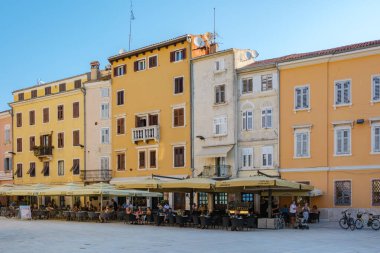 Rovinj Hırvatistan 27 Ağustos 2024, patronlar açık kafelerde sakin yemekler yiyip, güneş ve canlı atmosferi emmeyi seviyorlar. Büyüleyici sarı binalar meydanın resimli arkaplanına ekleniyor..