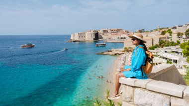 Bir gezgin, taş bir duvarda huzur içinde oturup Dubrovnik 'in ışıldayan turkuaz sularını seyrediyor. İnsanlar sıcak güneşin altında tarihi mimari ile çevrili plajın tadını çıkarırken tekneler sürükleniyor..