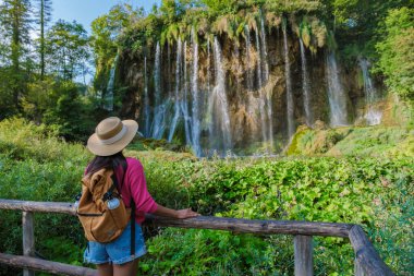 Bir gezgin Plitvice Gölleri 'ndeki kayalık uçurumlarda çağlayan şelalelere hayran, yemyeşil ve canlı bitki örtüsüyle çevrili. Güneş huzurlu atmosferi aydınlatıyor..