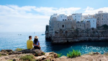 A couple relaxes on a rocky ledge, gazing at the stunning coastal landscape of Puglia, Italy, where whitewashed buildings meet the azure sea under a bright sky. Polignano a Mare Italy  clipart