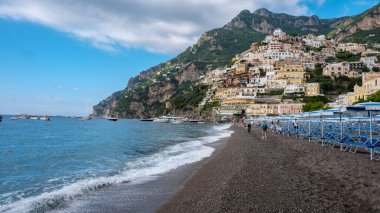 Ziyaretçiler, Positano 'nun kayalıklarından dökülen renkli evlerin nefes kesici manzarasının tadını çıkararak çakıllı sahillerde geziniyorlar. Ilık güneşin altında canlı mavi deniz parıldıyor, keşif yapmaya davet ediyor..