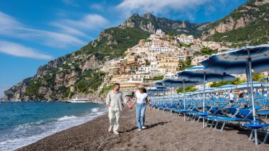 Bir çift, yamaçtaki renkli evlerle çevrili çakıllı Amalfi Sahili plajı boyunca romantik bir yürüyüş yapıyor. Gökyüzü açık ve mavidir, deniz kenarında unutulmaz bir gün için mükemmeldir..