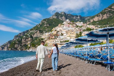 Bir çift, İtalya, Positano 'da, sıcak güneşin altında, renkli uçurum kenarındaki evler ve kıyıya vuran nazik dalgalarla el ele yürür..