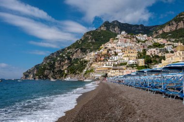 Amalfi kıyısında, Positano 'nun pitoresk kasabası kayalıklardan dökülen renkli binalar, parlak bir gökyüzü altında kumlu plaj sandalyeleri ve hafif dalgalarla kaplı..