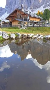 Büyüleyici bir ahşap kulübe kristal berrak bir göletin yanında huzur içinde duruyor, çarpıcı dağları ve canlı sonbahar renklerini yansıtıyor. Geisler Alm Odle Dağı, Alto Adige, Dolomitler, İtalya