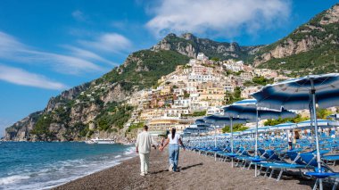 Bir çift, İtalya 'nın Positano kentindeki kumlu sahil boyunca yürüyor. Uçurumların üzerinde renkli evler var ve parlak bir gökyüzünün altında mavi şemsiyeler var..