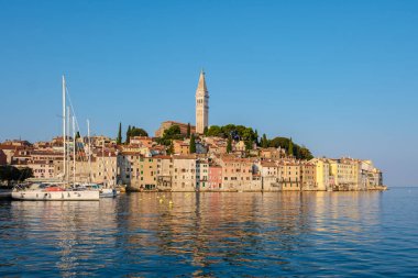 Tarihi mimarinin açık mavi gökyüzüyle buluştuğu Rovinj 'in canlı kıyı şeridine hafif dalgalar vuruyor. Resimli bir yelkenli gemi güneş doğarken demirli duruyor..