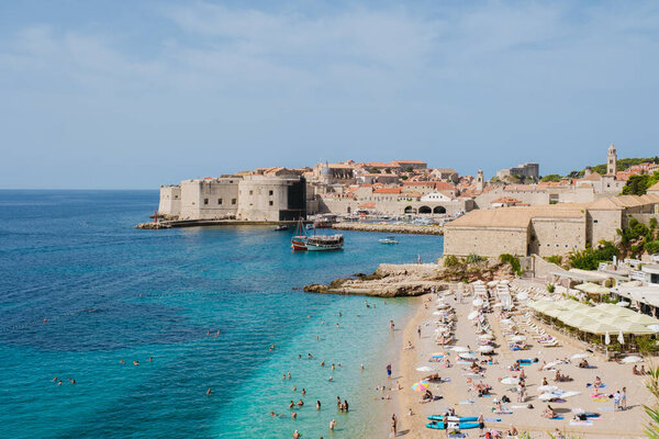 Посетители отдыхают на песчаном пляже под теплыми солнечными лучами, наслаждаясь чистой бирюзовой водой и потрясающим видом на древние укрепления Дубровника и очаровательную гавань.
